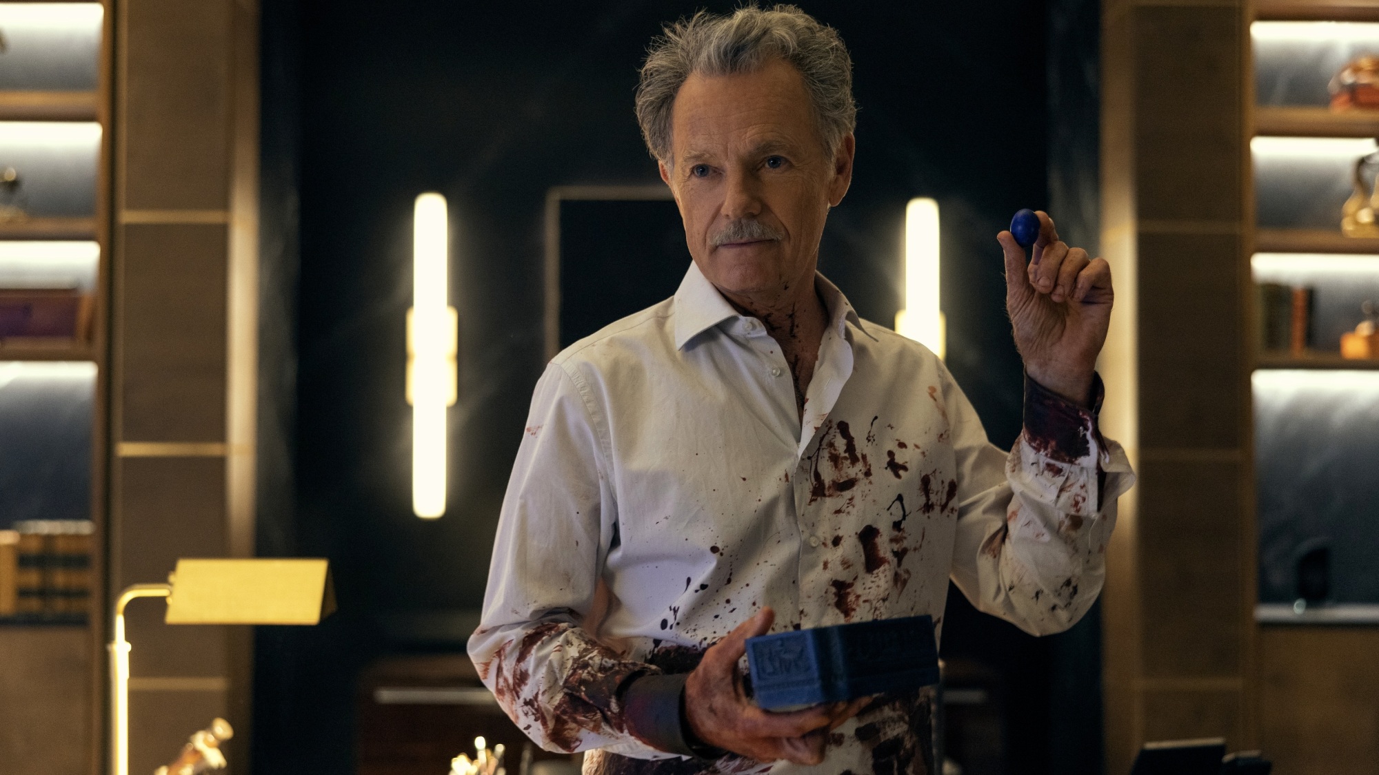 A man in a bloody white dress shirt holds a blue box and a polished blue sapphire.