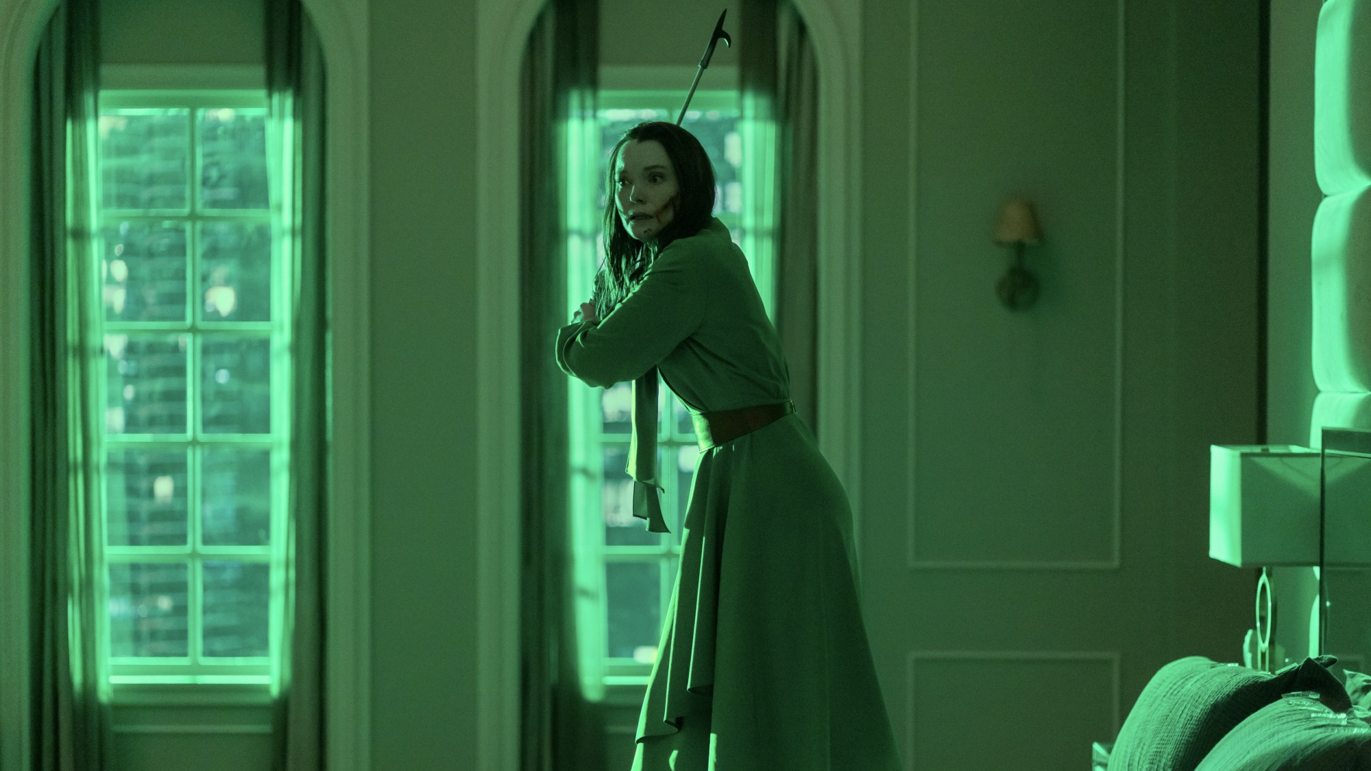 A woman in a green dress stands on a bed in a green-lit room, holding a fireplace poker like a weapon.