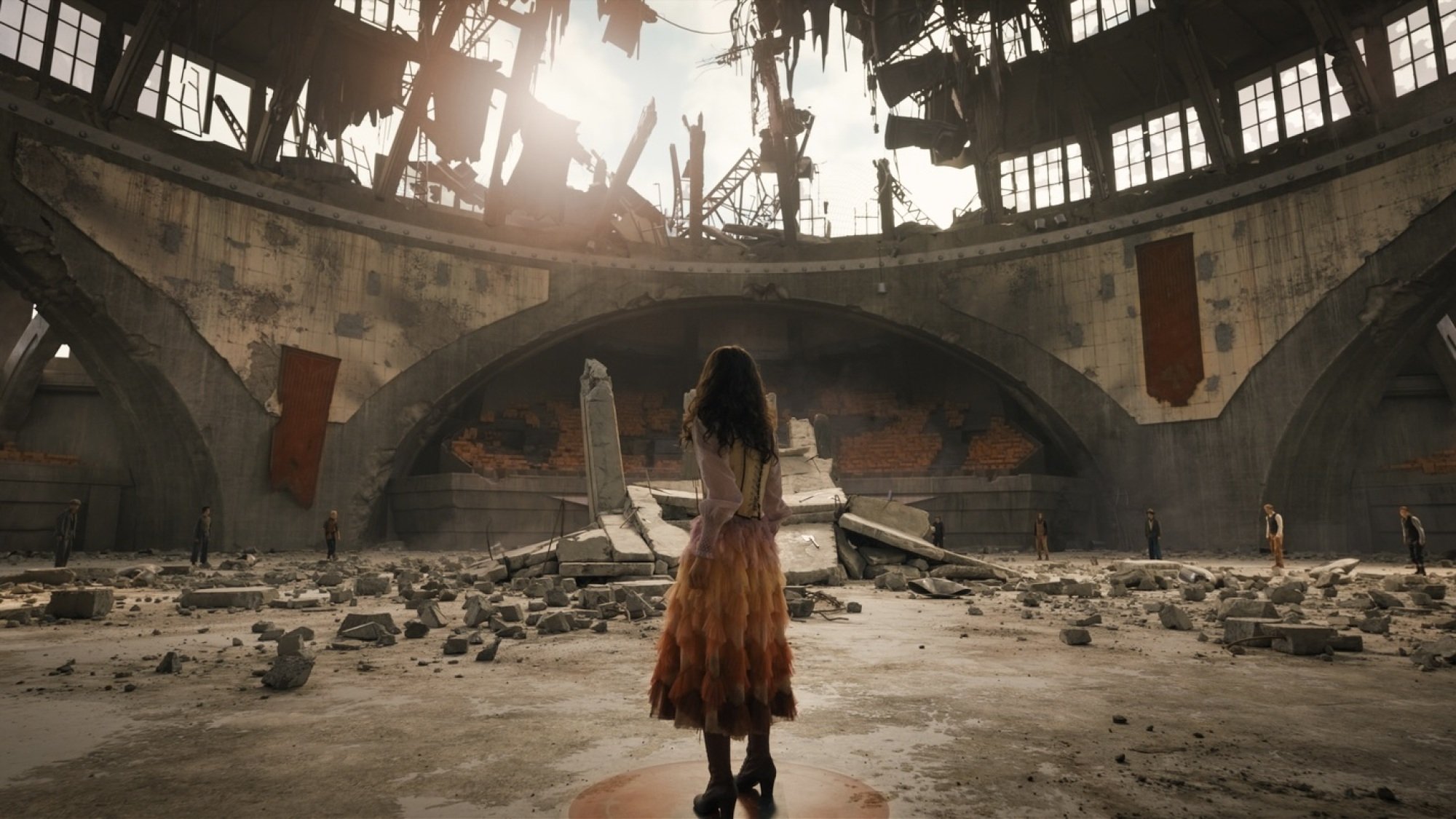 A young woman in a colorful dress stands in a ruined arena.
