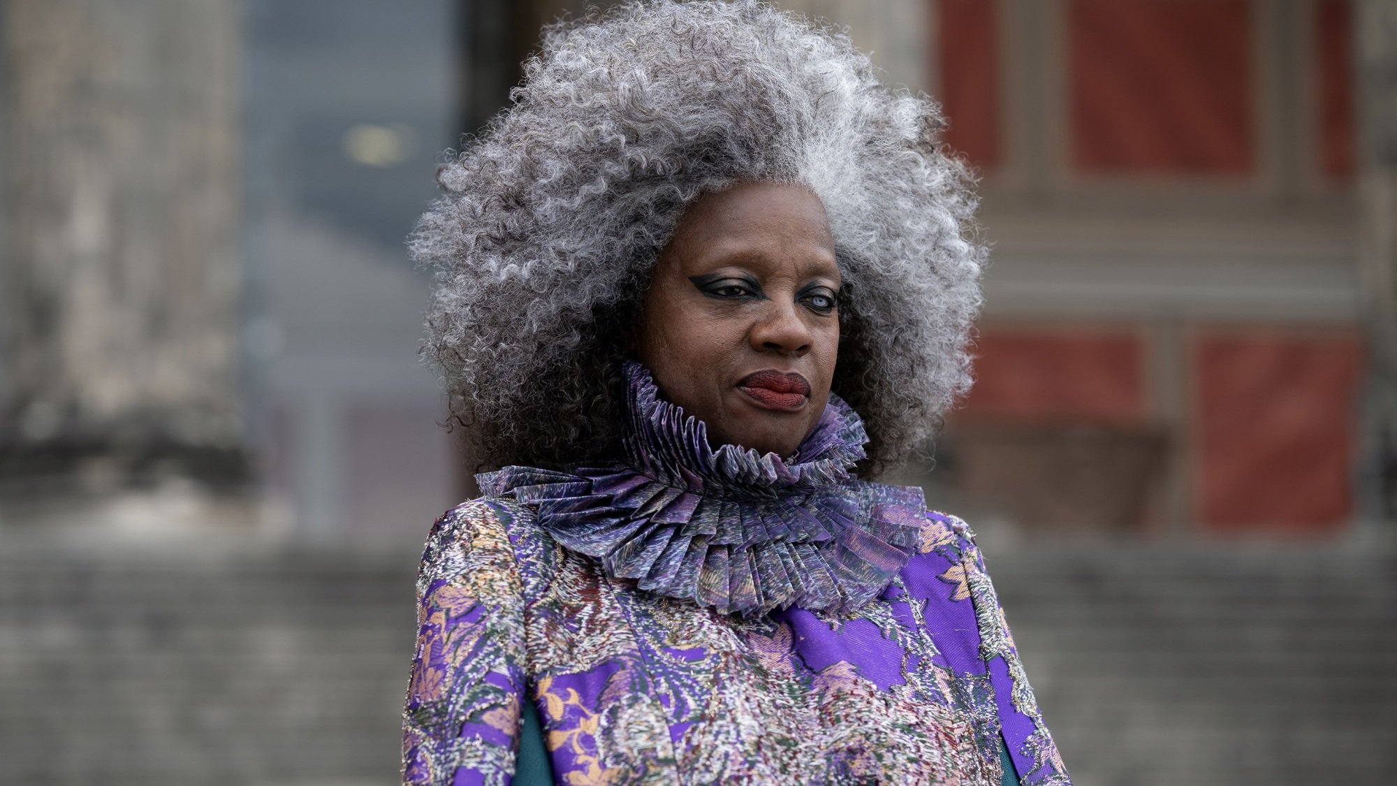 A woman with greying hair, one brown eye, and one milky blue eye wearing an elaborate purple dress.