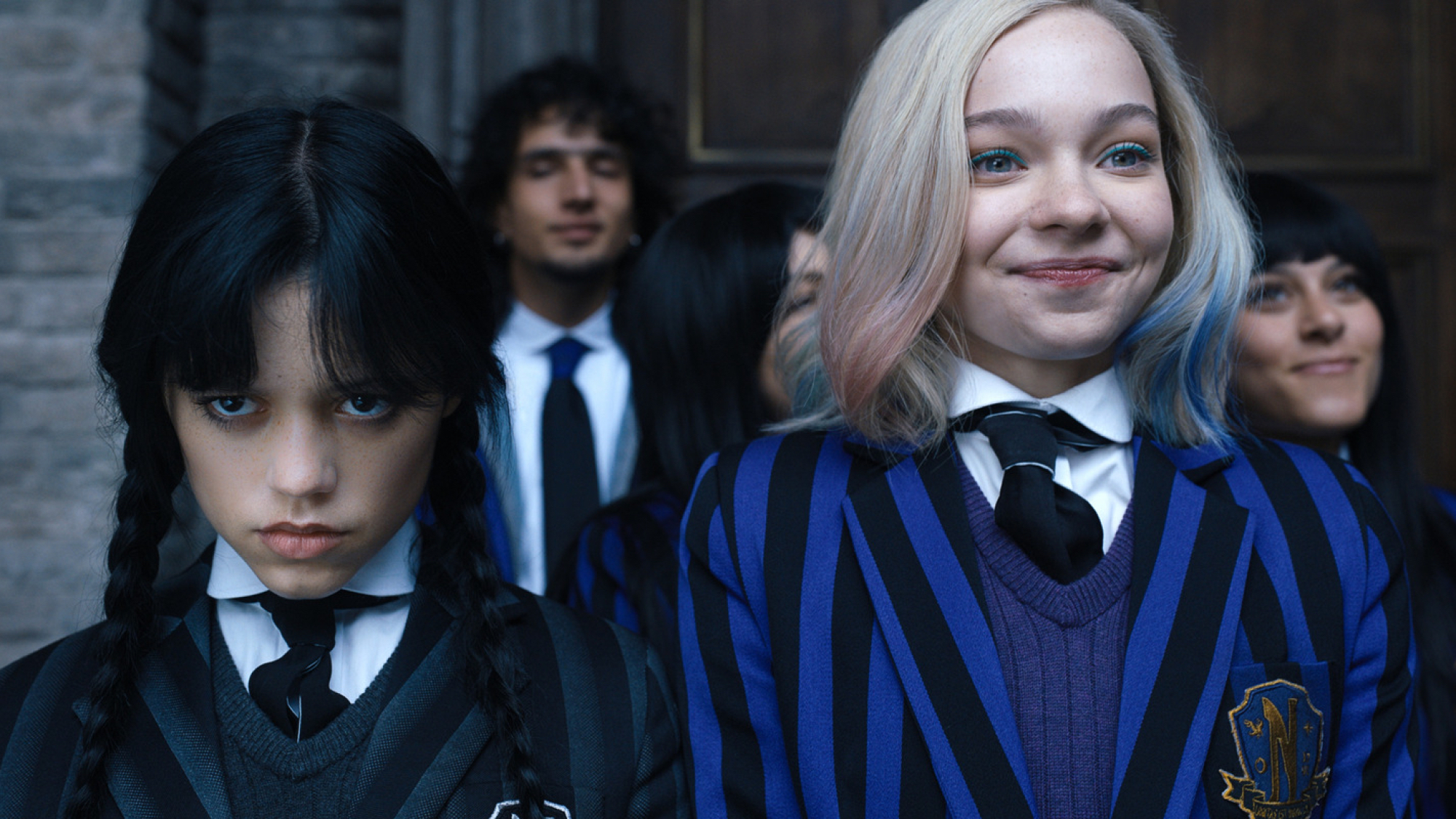 Two teen girls stand next to each other in school uniform, one smiling while the other scowls.