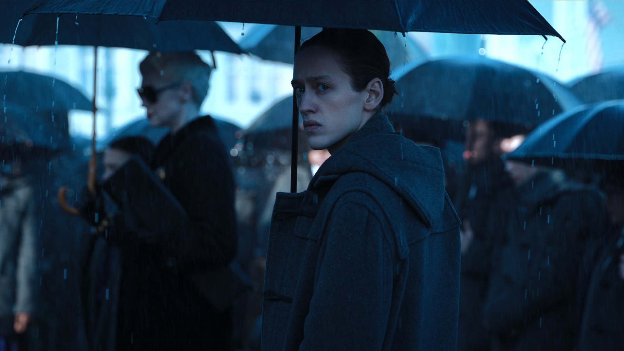 A teen boy stands at a funeral with an umbrella in the rain.