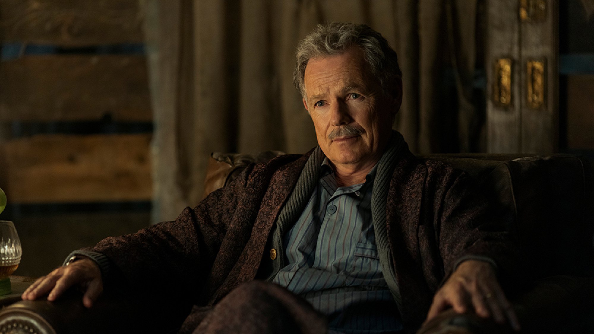 An older man sits in an armchair in a firelit room.