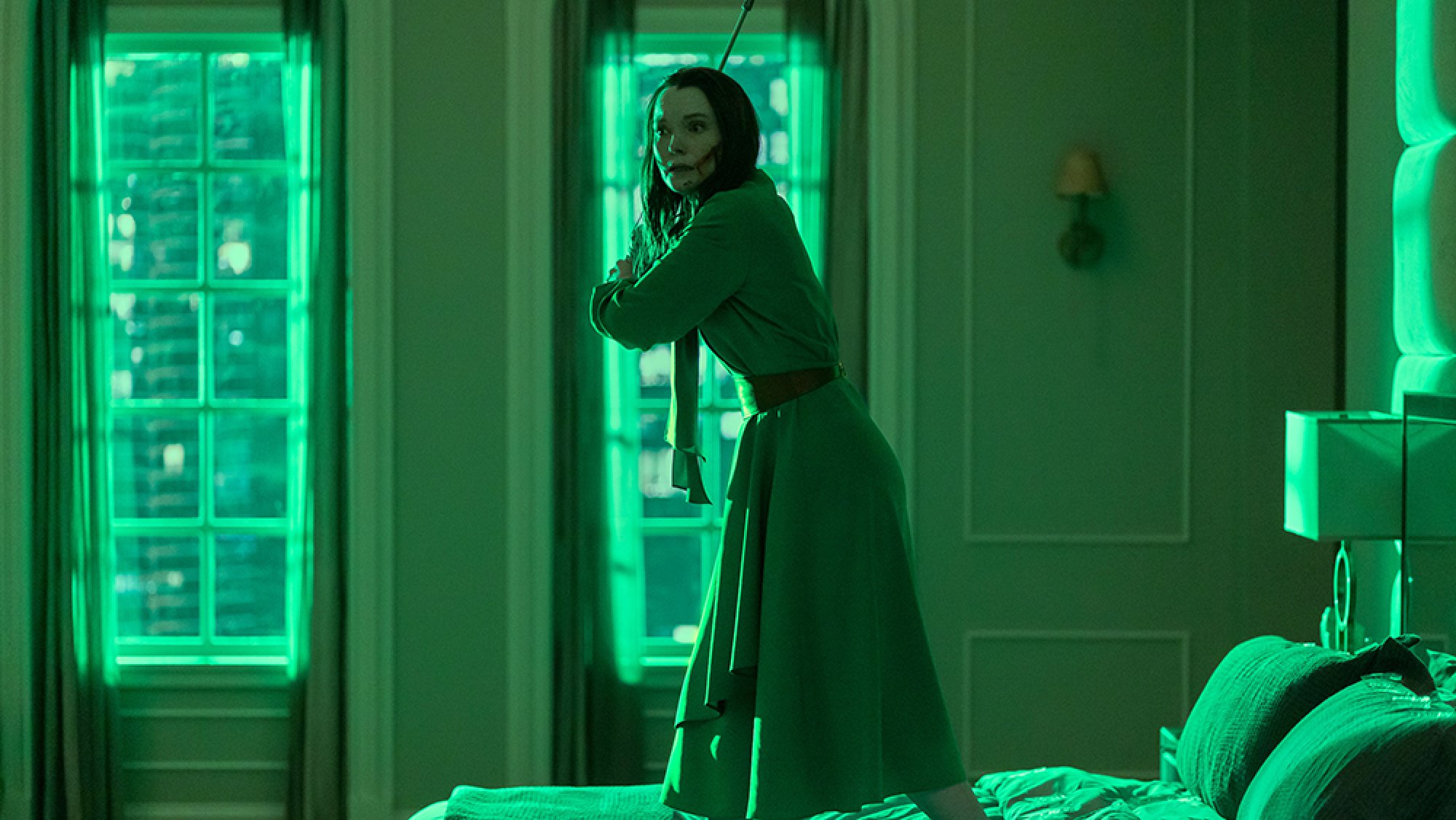 A woman stands on a bed in a green-lit room, wielding a fire poker.