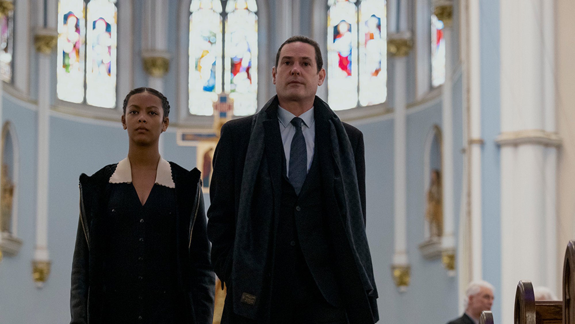A man in a suit walks through a church, while a teenage girl follows behind him.