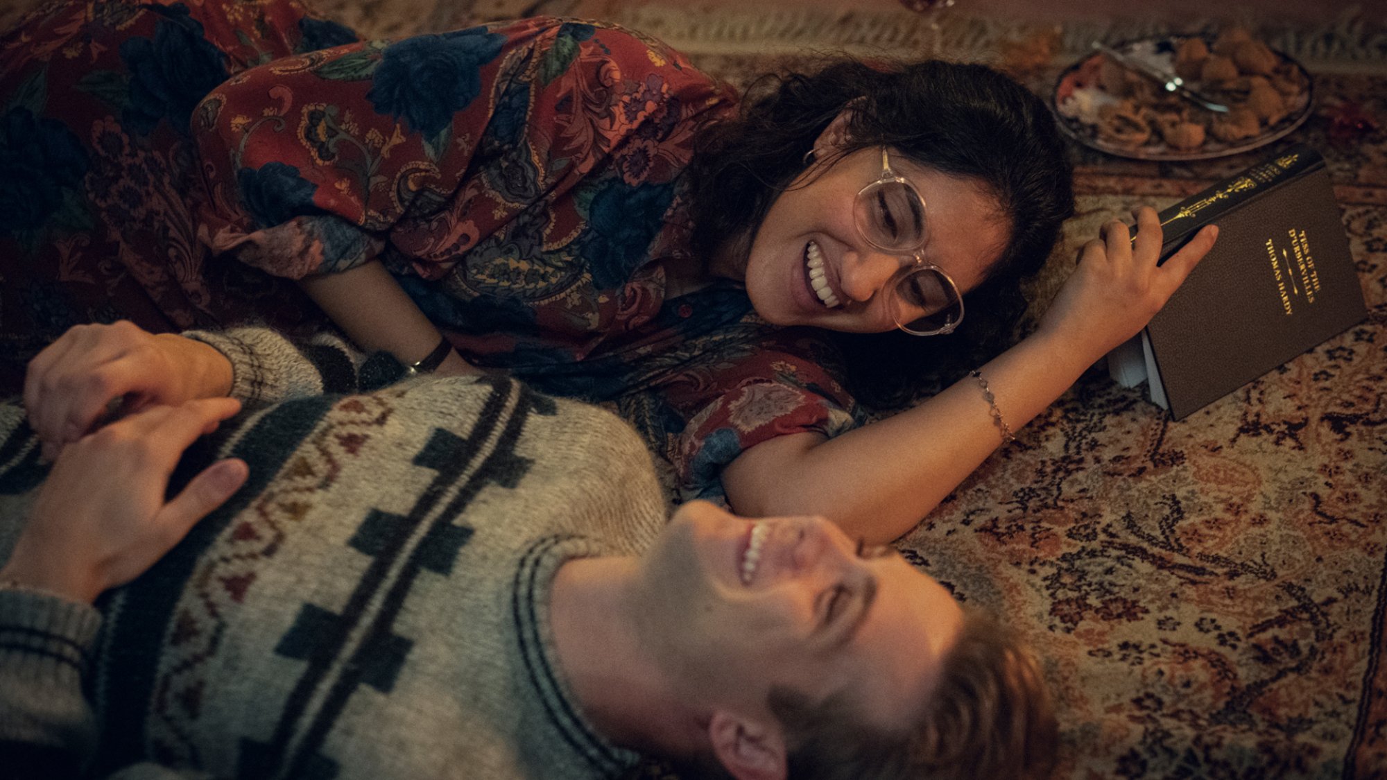 A young woman and a young man lie laughing on a carpet reading a book.