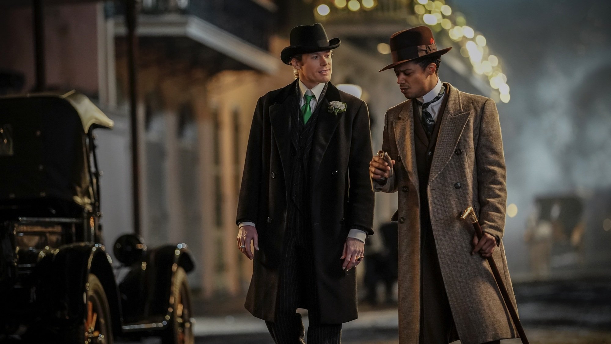 Two men in suits, long coats, and hats walk down a New Orleans street.