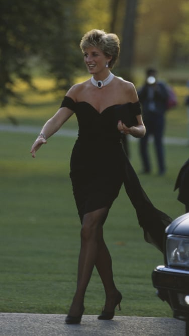 A woman in an off-the-shoulder black evening gown walks holding a clutch.