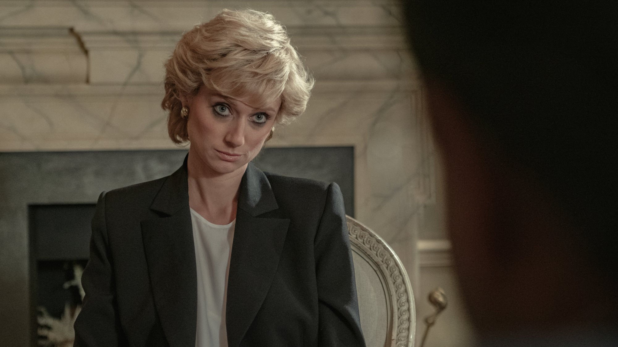A woman with blonde short hair wearing a black blazer sits opposite an interviewer.