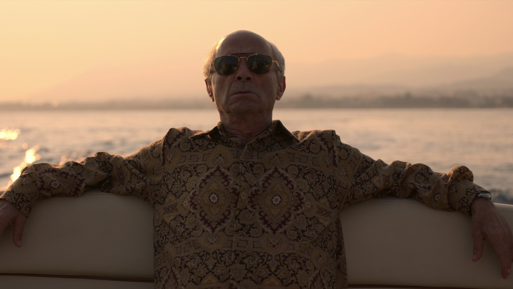 A man in a patterned shirt wearing sunglasses reclines at the back of a boat.
