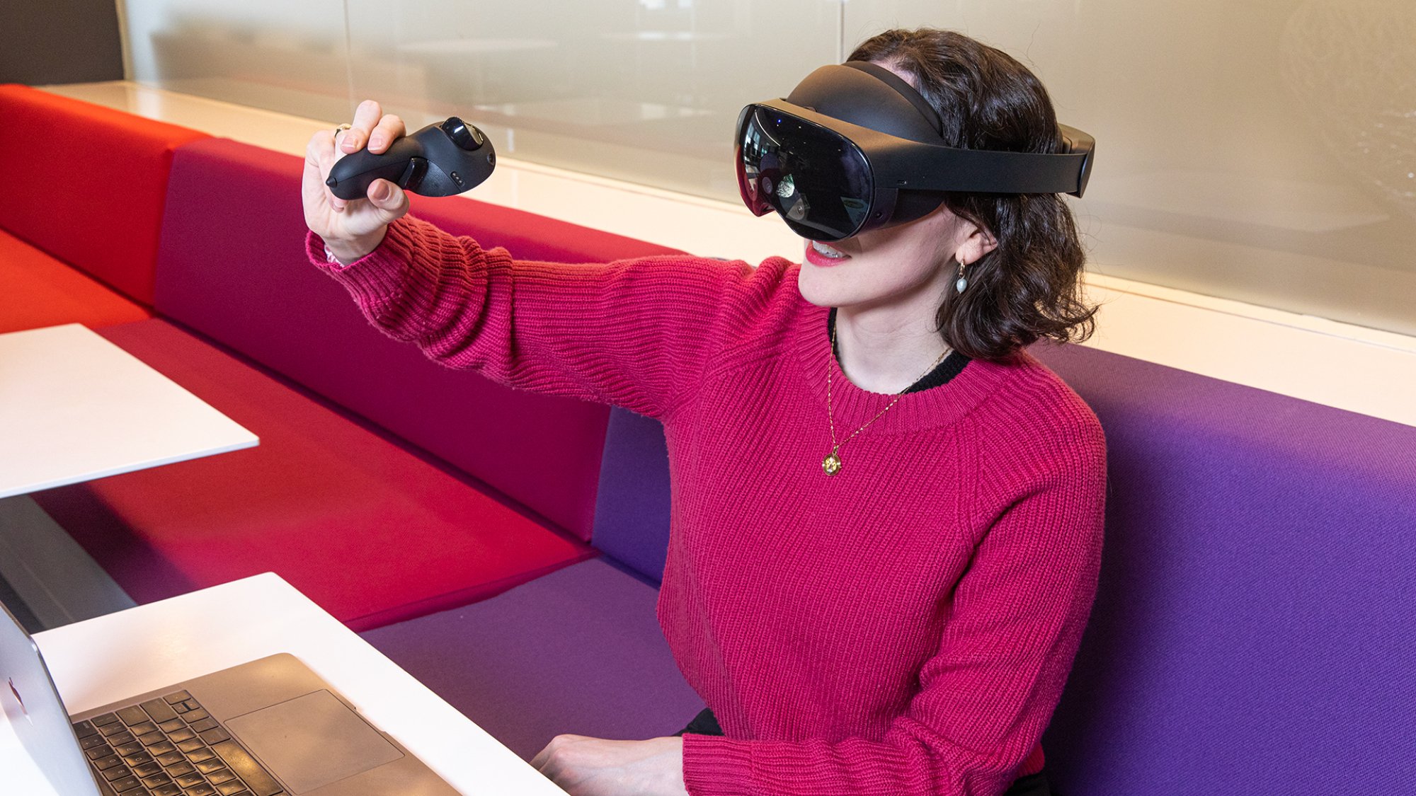 A woman sits at a purple booth in front of a laptop. She has short brown hair and a fuchsia sweater. She is wearing the Quest Pro and holding one of its controllers, which has been fitted with a stylus at the bottom, and is drawing in the air.