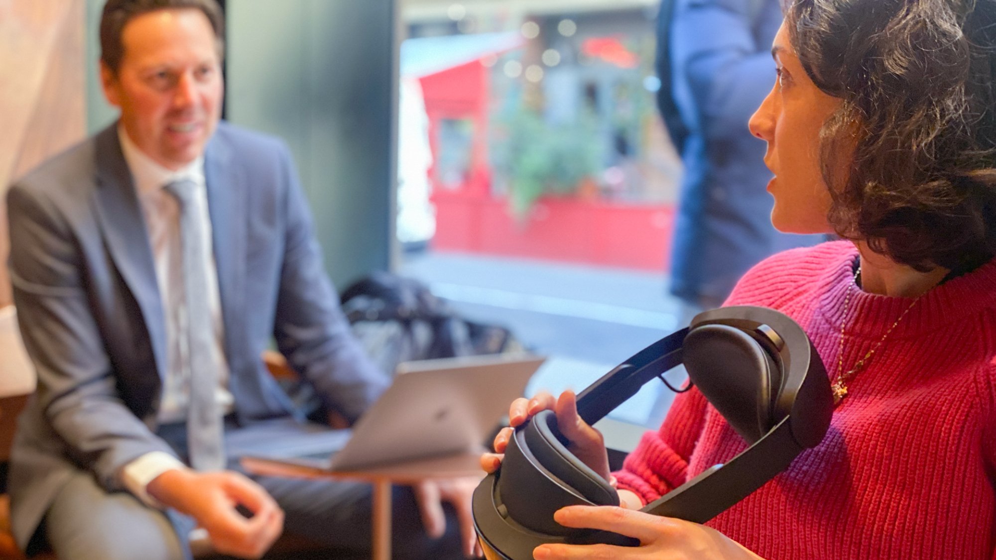 A cafe. In the foreground, a woman holds a Meta Quest Pro and has her head turned in profile. She is talking to a white man in the background who is dressed in a business suit.
